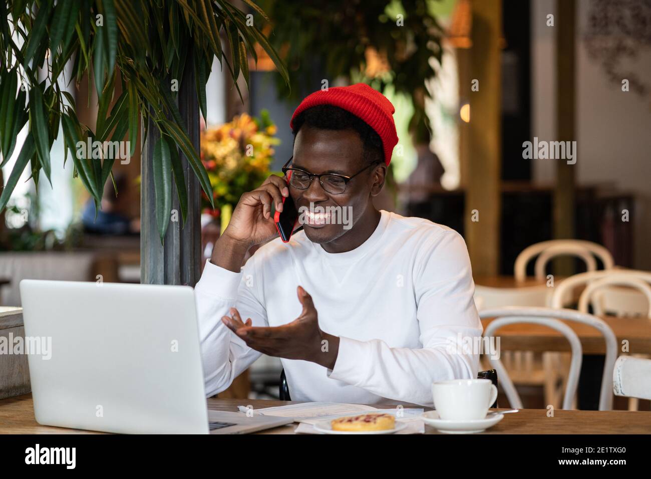 Happy millennial Black man laughing, speaking with friend on mobile phone, remote work in cafe. Stock Photo