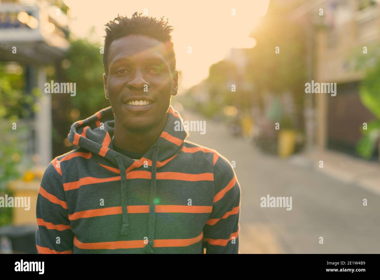 Young happy slim black African man smiling with sunlight beaming at the back outdoors Stock Photo