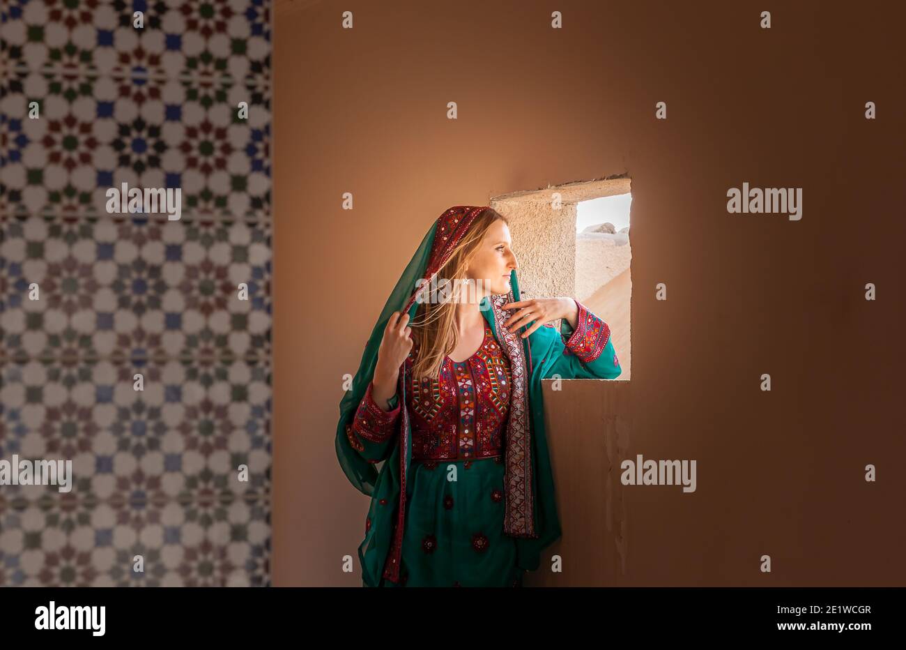 gorgeous shy girl in traditional omani clothes looking through the window and covering her face with veil to hide from eyes of people walking nearby Stock Photo