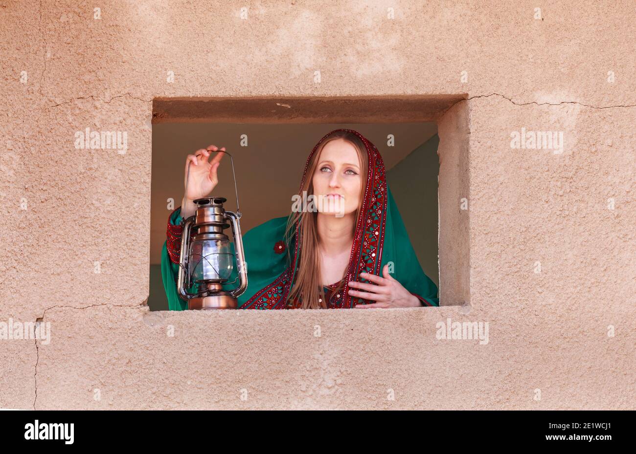 gorgeous shy girl in traditional omani clothes looking through the window and covering her face with veil to hide from eyes of people walking nearby Stock Photo