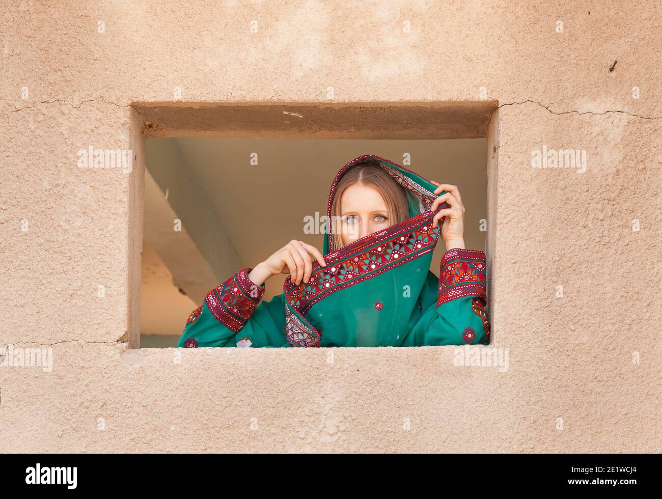 gorgeous shy girl in traditional omani clothes looking through the window and covering her face with veil to hide from eyes of people walking nearby Stock Photo