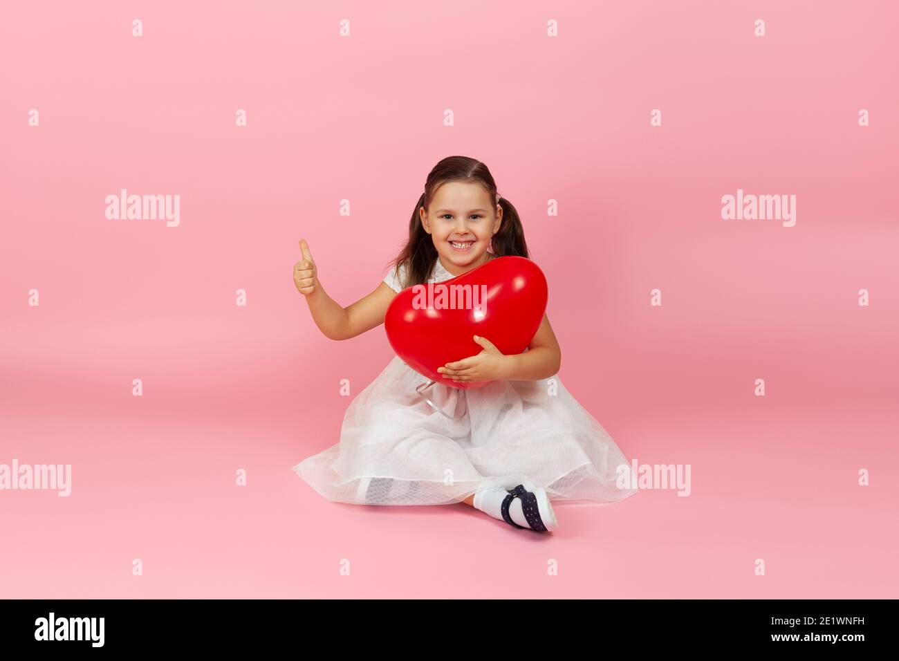 attractive, gorgeous girl in white dress holding red heart shaped balloon and sitting on the floor and giving thumbs up isolated on a pink background Stock Photo