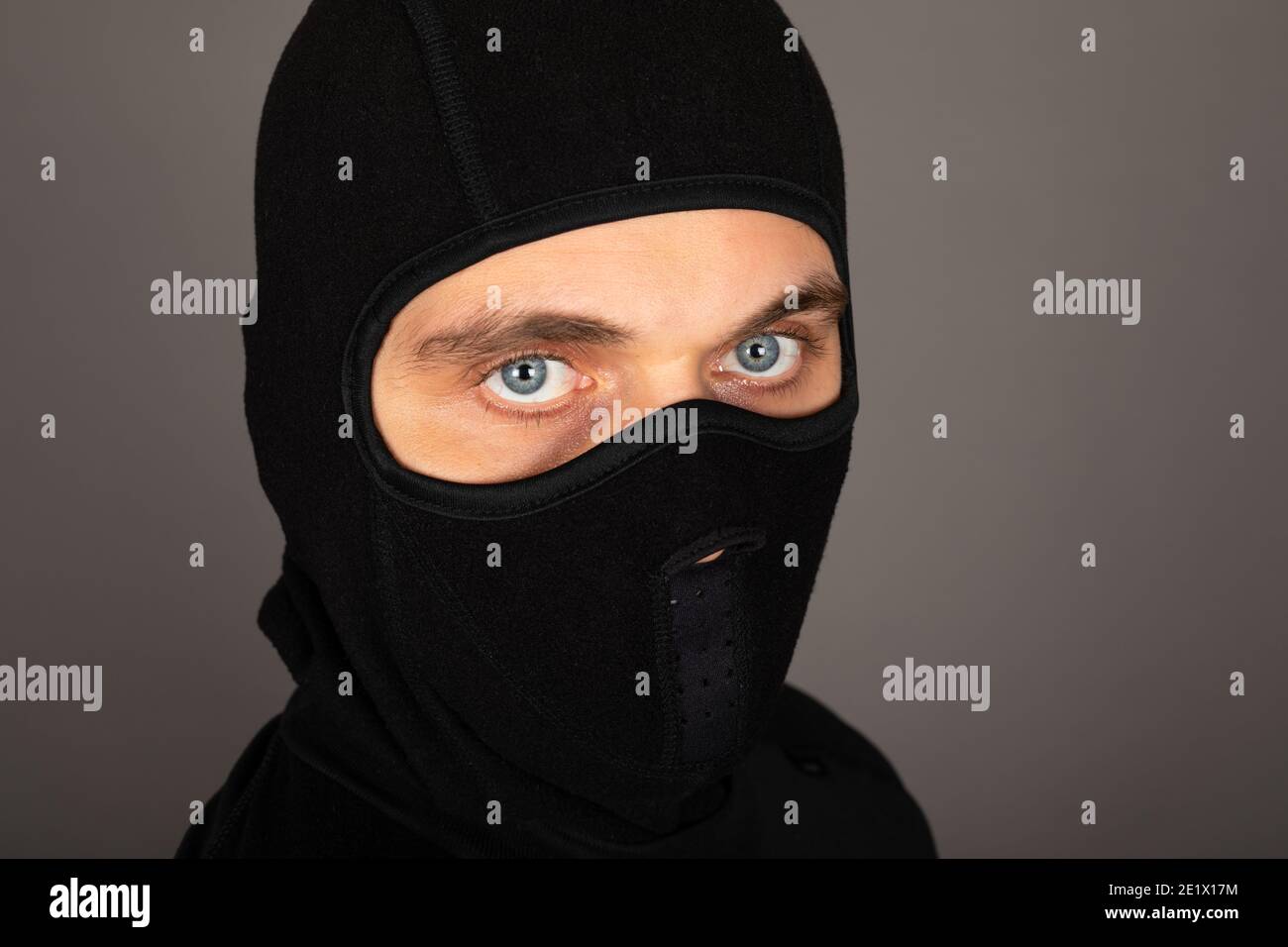 Picture of young man with black mask and outfit suspect of a robbery, wearing handcuffs in front of grey background Stock Photo