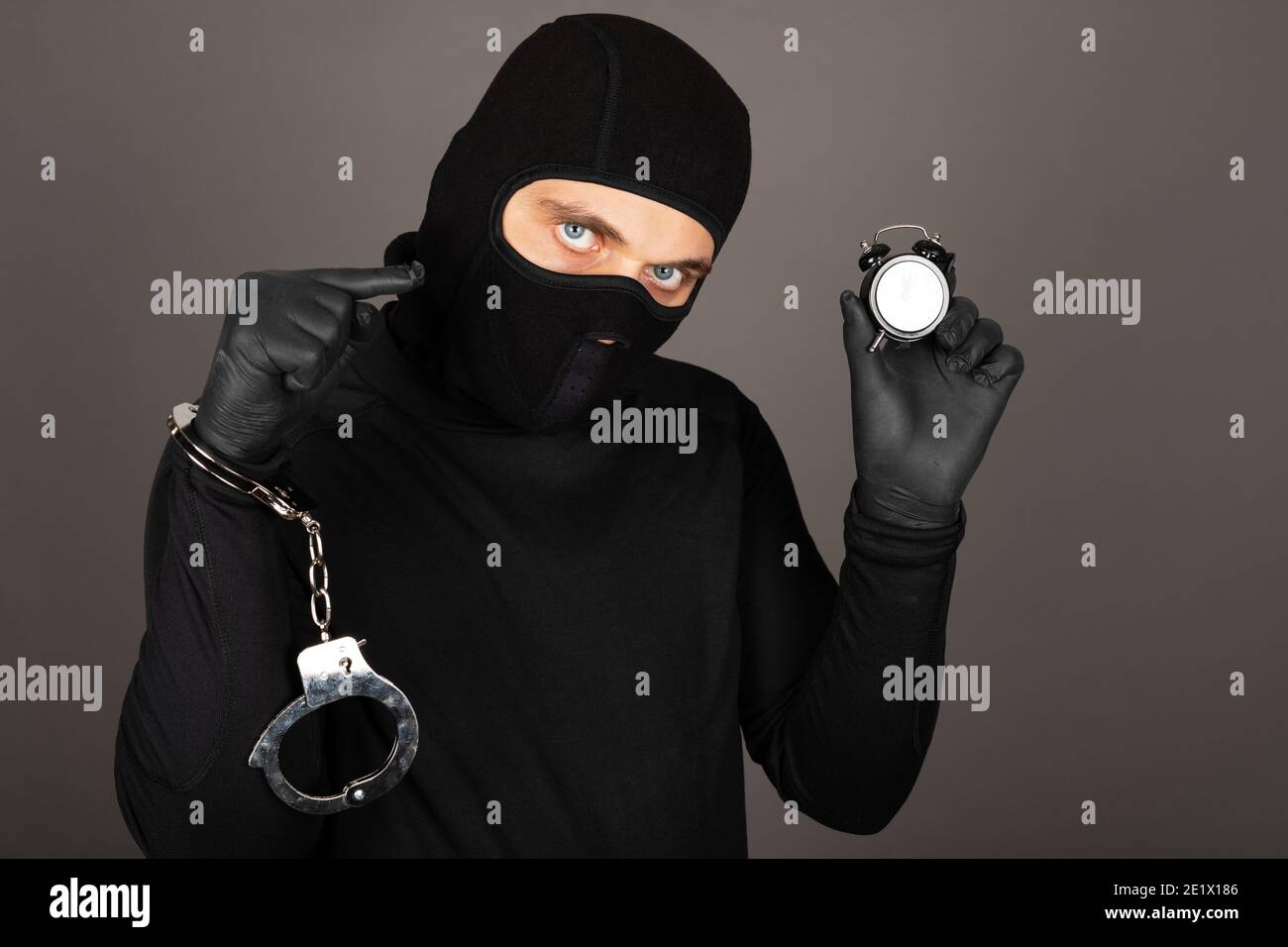 Picture of young man with black mask and outfit suspect of a robbery, wearing handcuffs in front of grey background Stock Photo