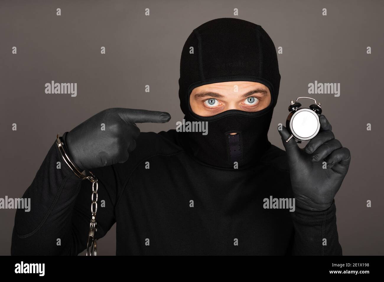 Picture of young man with black mask and outfit suspect of a robbery, wearing handcuffs in front of grey background Stock Photo