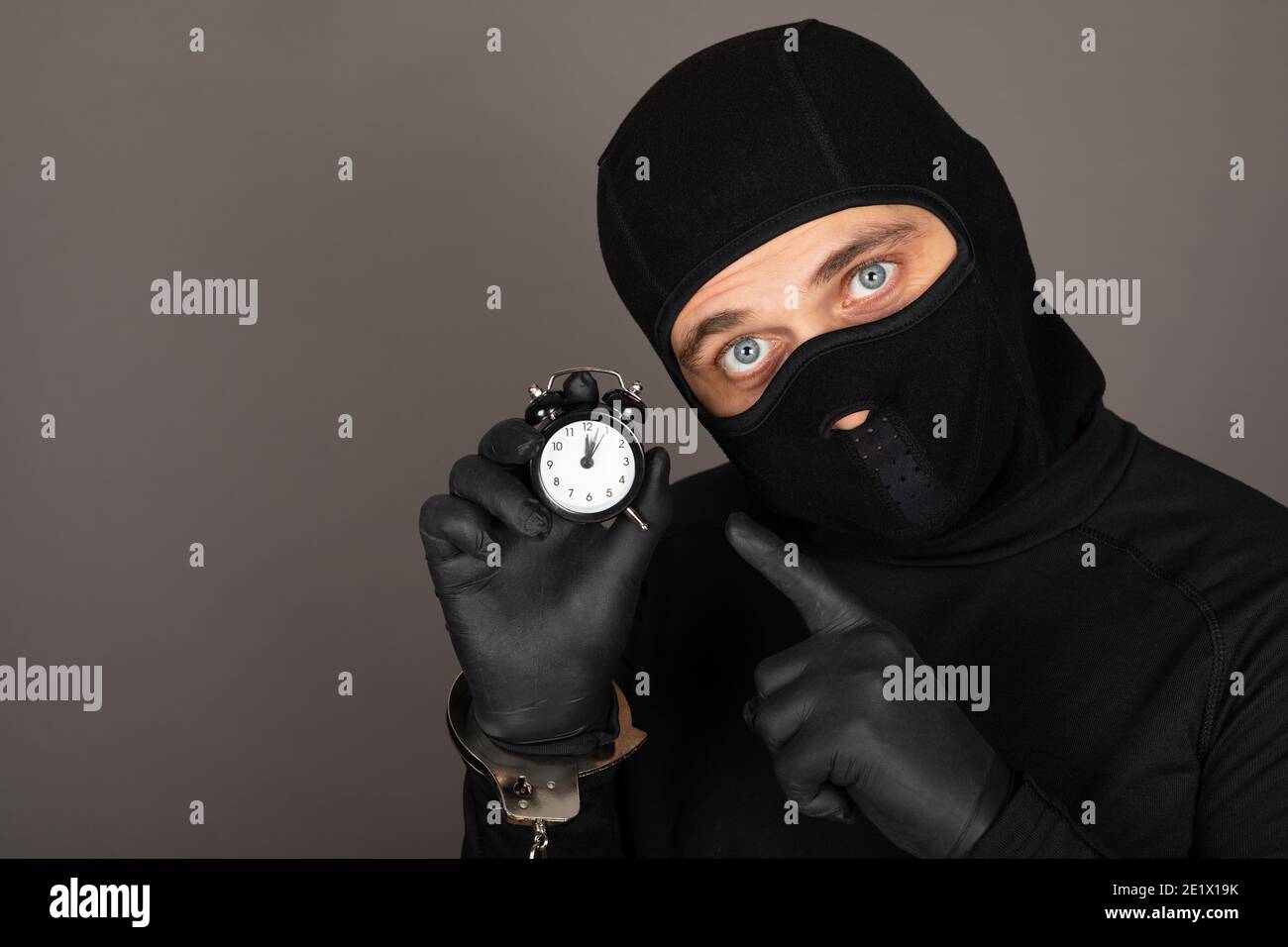 Picture of young man with black mask and outfit suspect of a robbery, wearing handcuffs in front of grey background Stock Photo