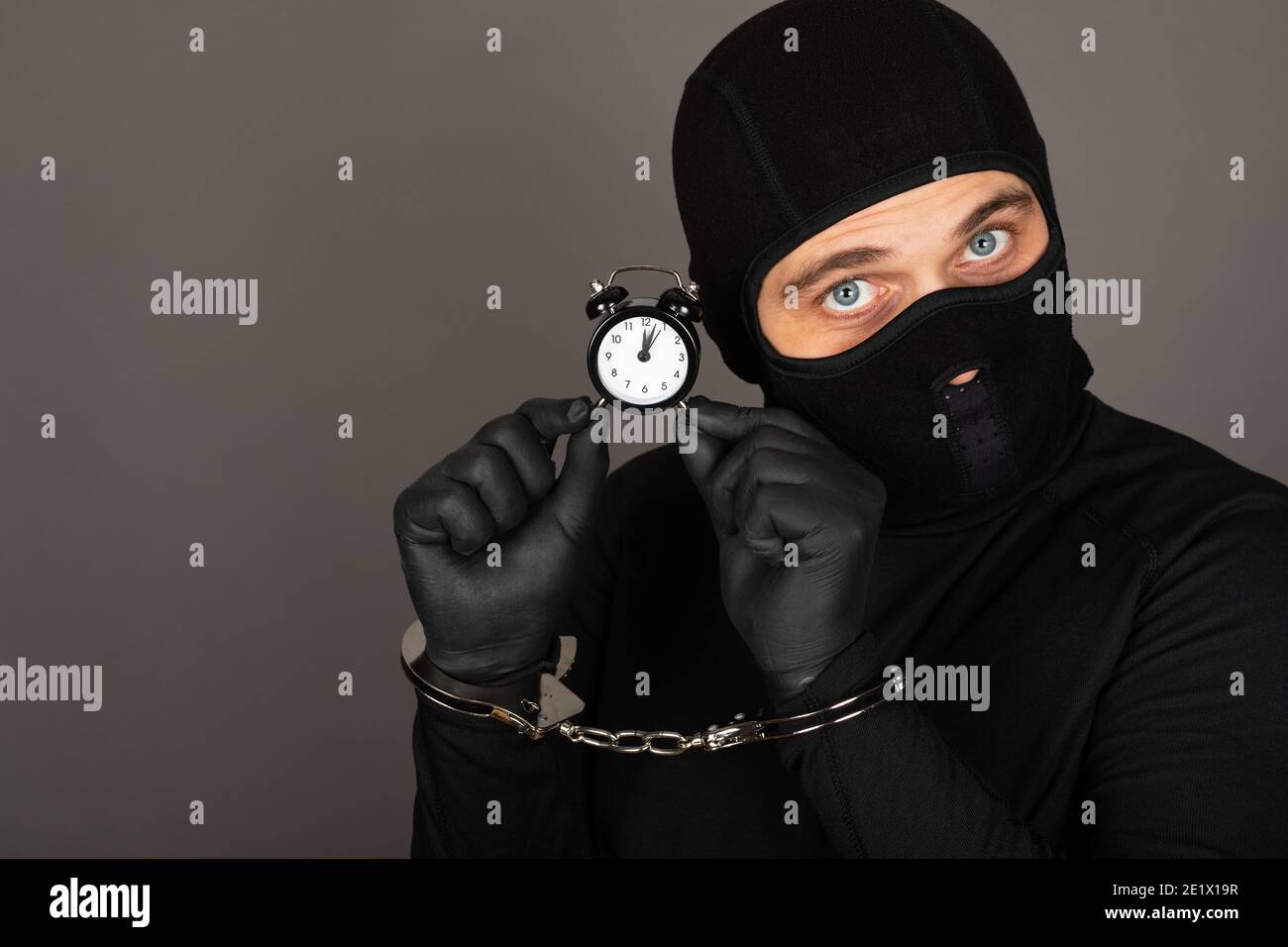 Picture of young man with black mask and outfit suspect of a robbery, wearing handcuffs in front of grey background Stock Photo