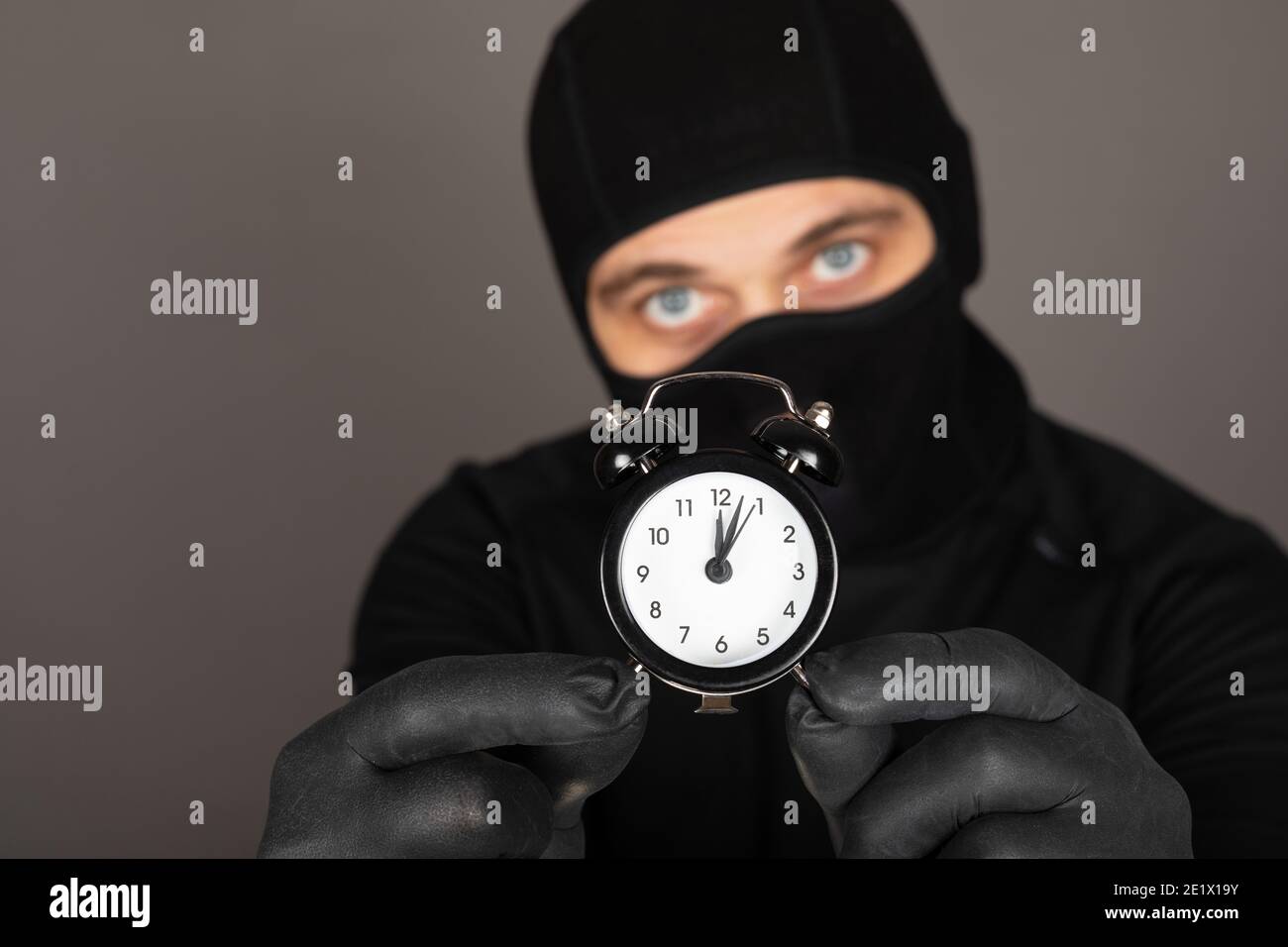 Picture of young man with black mask and outfit suspect of a robbery, wearing handcuffs in front of grey background Stock Photo