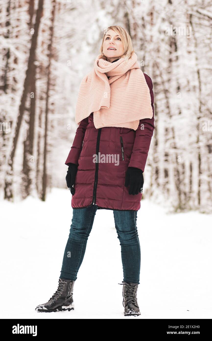 Portrait of blonde woman with purple coat and pink scarf standing in the forest during winter time and watching nature Stock Photo
