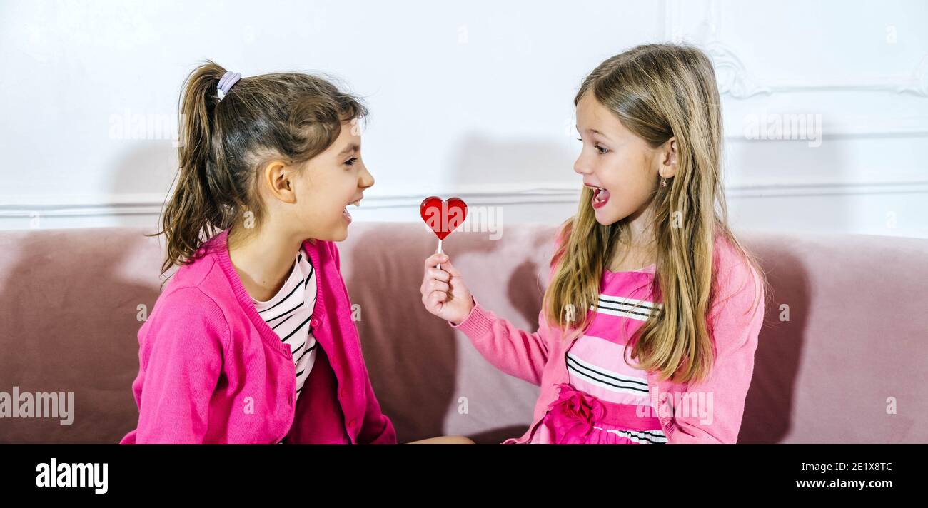 Close-up of two happy little girls with heart shape lollipop standing together at home. Valentines day kids friendship concept Stock Photo