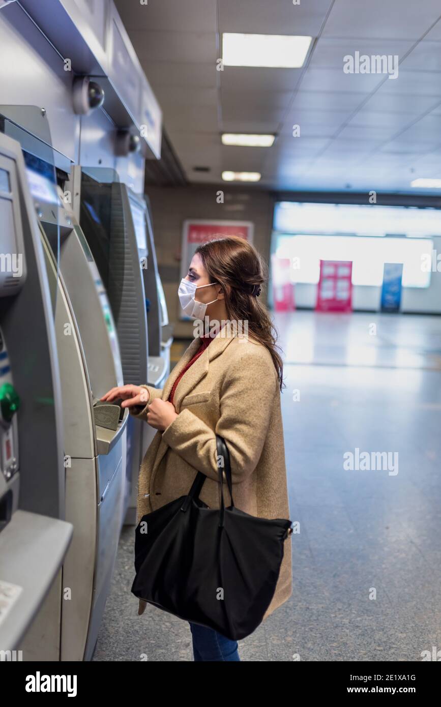 Beautiful girl wearing protective medical mask and fashionable clothes uses ATM machine for money.New normal concept during pandemic corona,covid19. Stock Photo