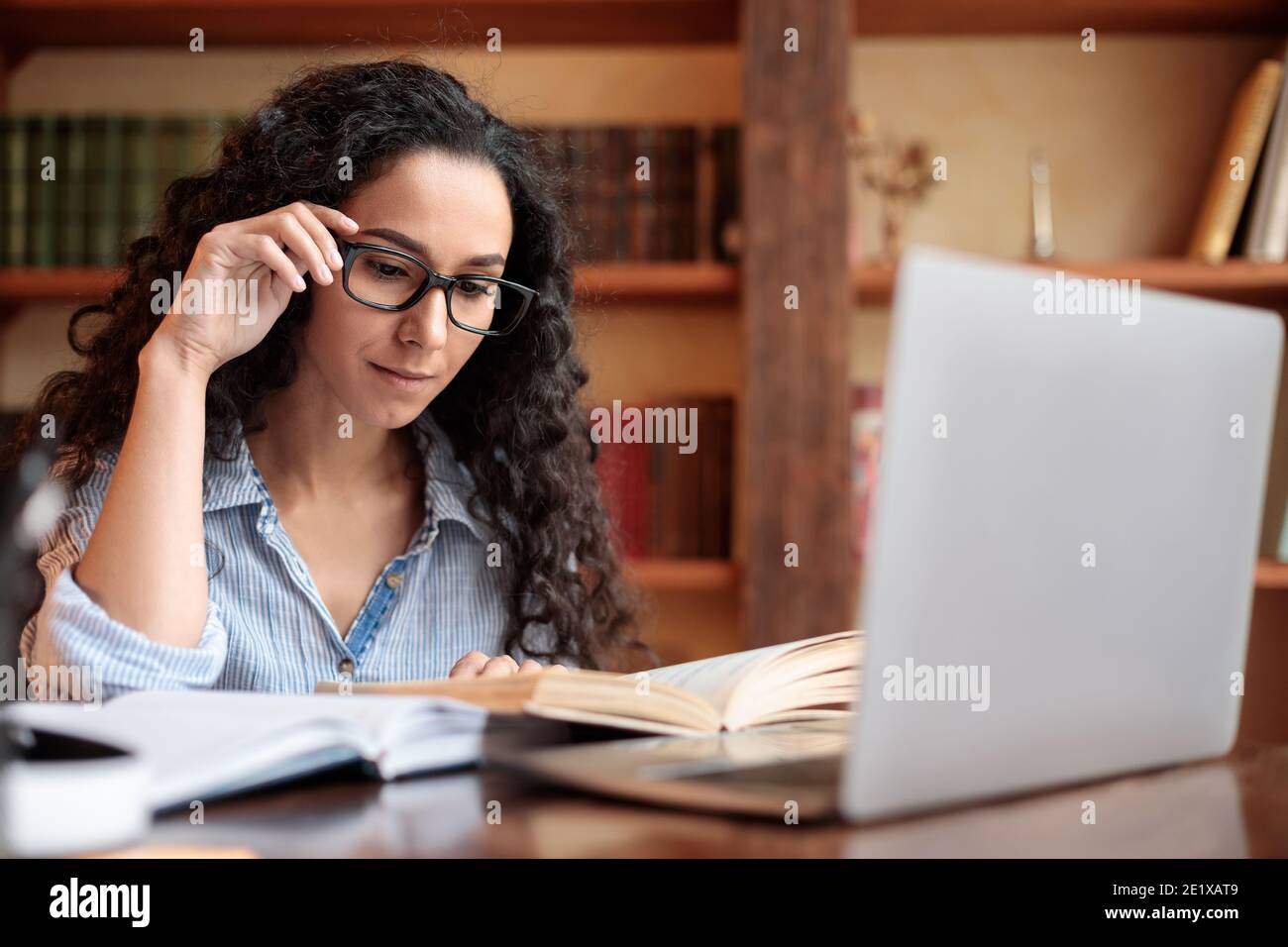 Young lady reading book sitting at desk touching glasses Stock Photo