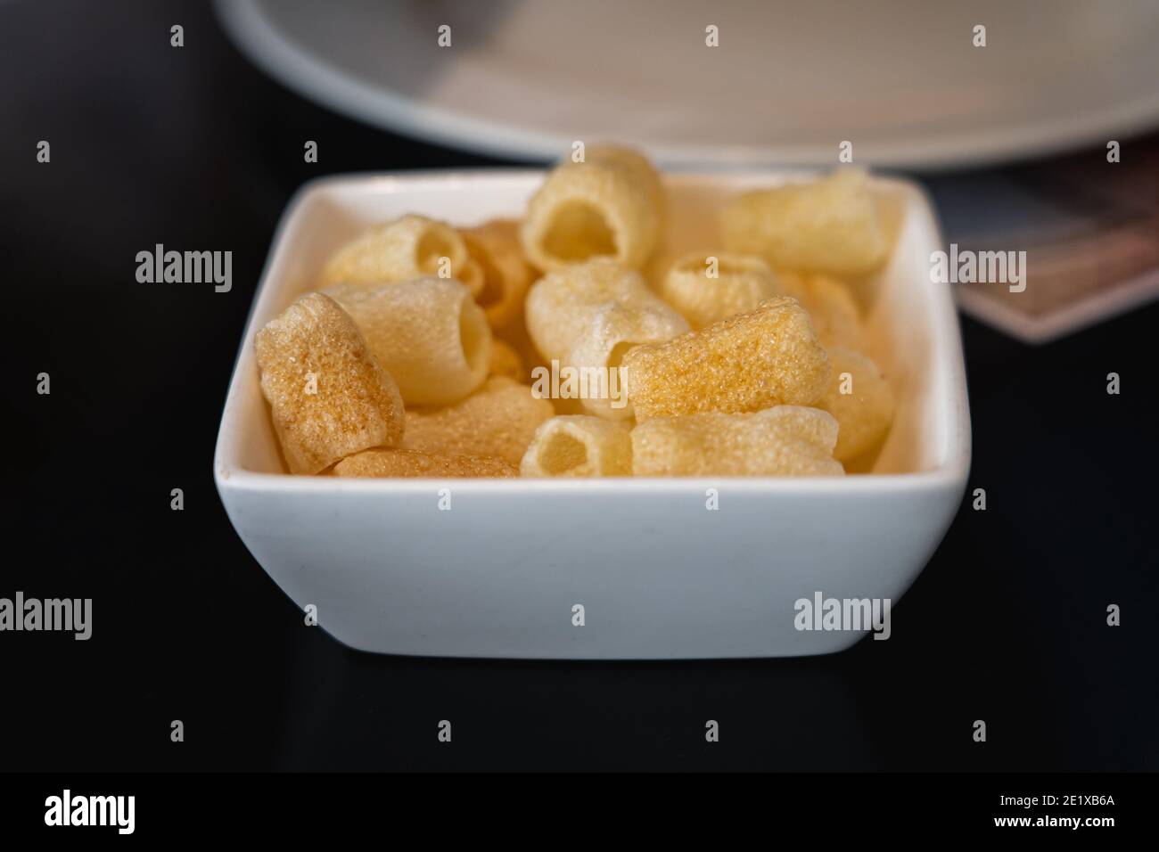 Fryums served in a small white bowl on a black table. A white plate in the background. Stock Photo