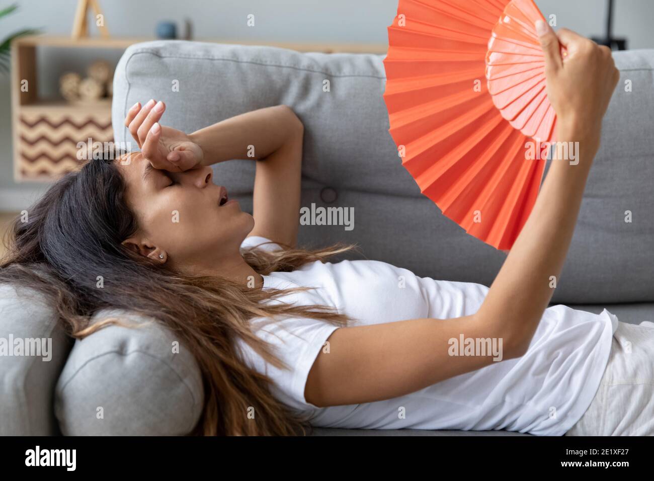 Close up overheated woman waving paper fan, lying on couch Stock Photo