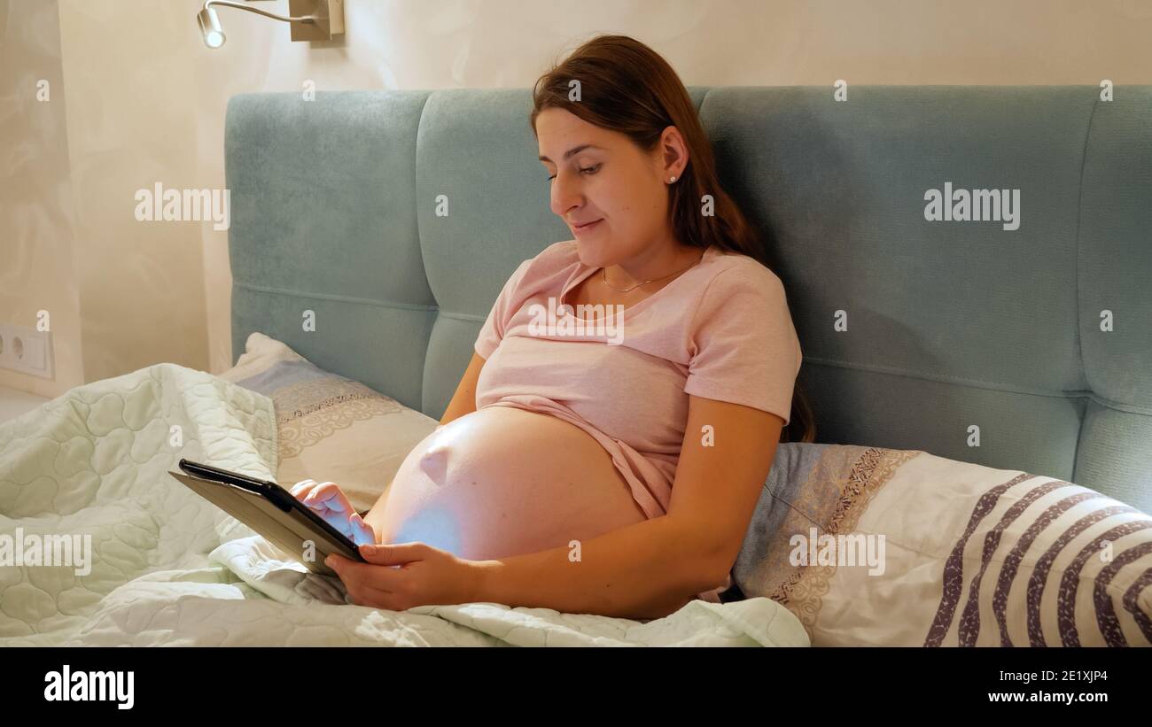 Smiling pregnant woman wearing pajamas working on tablet and browsing internet before going to sleep in bed at night Stock Photo