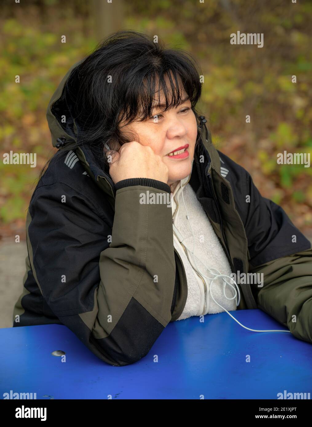 An Asian middle aged woman with in-ear headphones speaking in telephone. Blurry green nature background Stock Photo