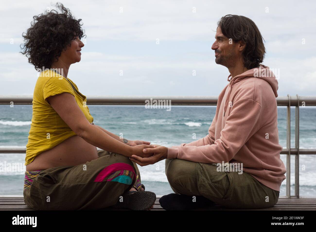 Expecting parents holding hands sitting on lotus pose on bench by beach in Las Palmas, Spain. Pregnant woman and future father enyoing leisure time Stock Photo