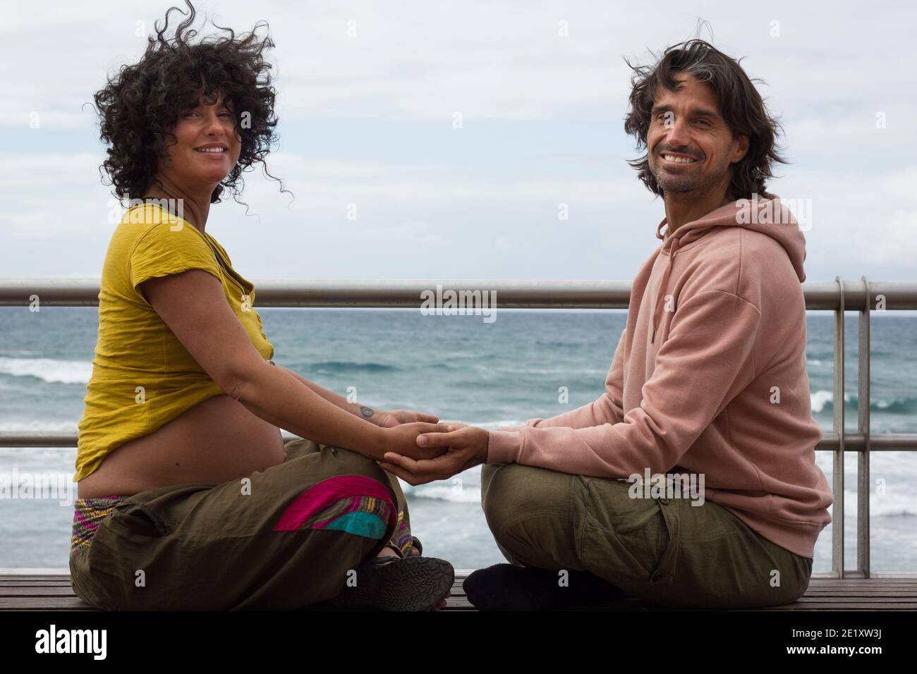 Smiling couple holding hands sitting on lotus pose on bench by beach in Gran Canaria, Spain. Pregnant woman and future father enyoing leisure time Stock Photo