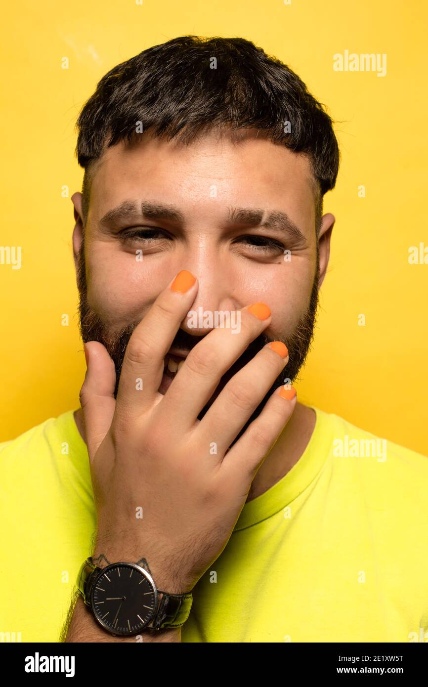Portrait of a young man with yellow beard and t-shirt on yellow background smiling and covering his mouth with hand with orange nail polish and black Stock Photo