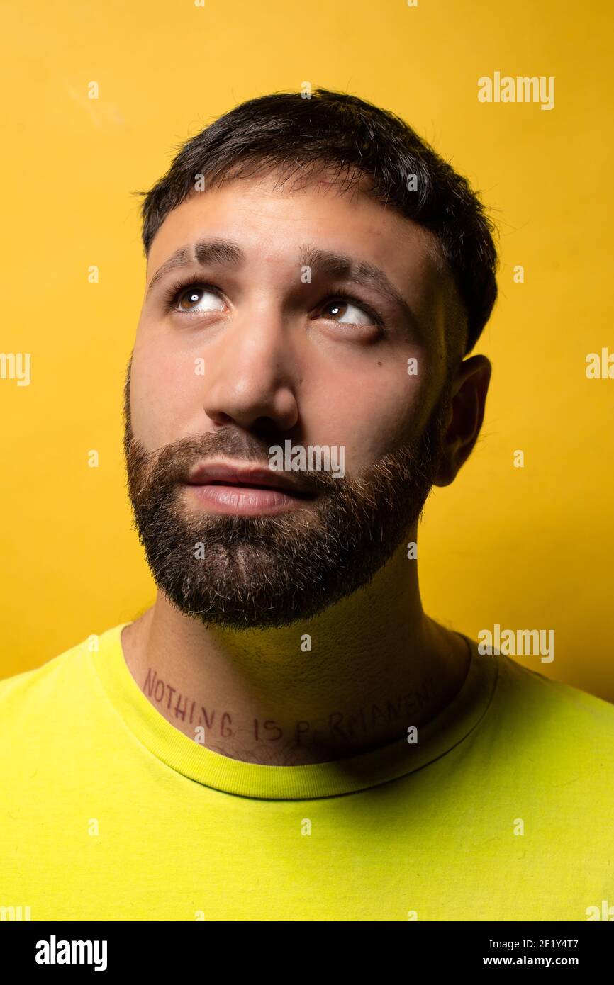 Portrait of a young man with beard and yellow t-shirt on yellow background looking up smiling Stock Photo