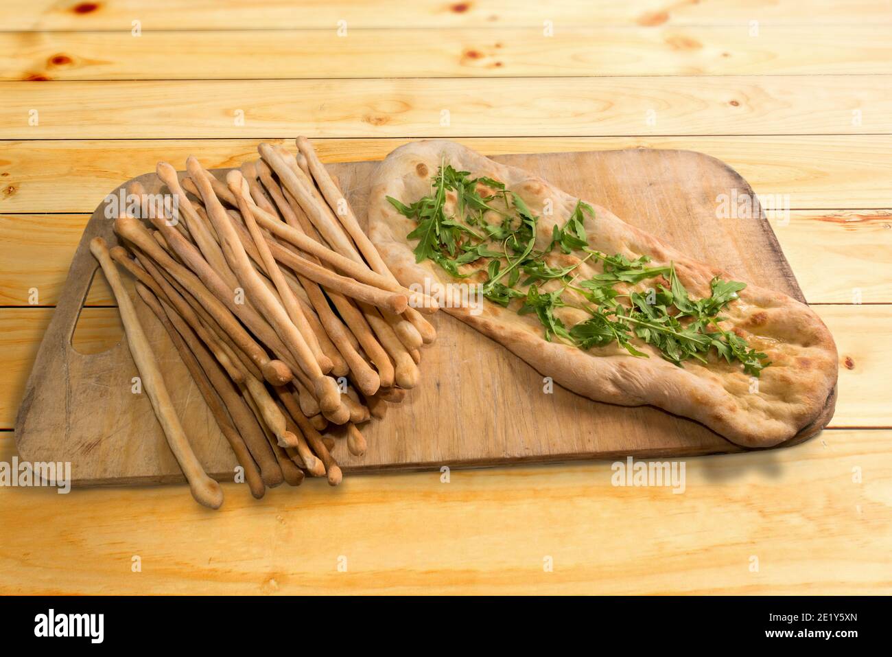 bread sticks and crushed focaccia with oil with rocket leaves on wooden cutting board Stock Photo