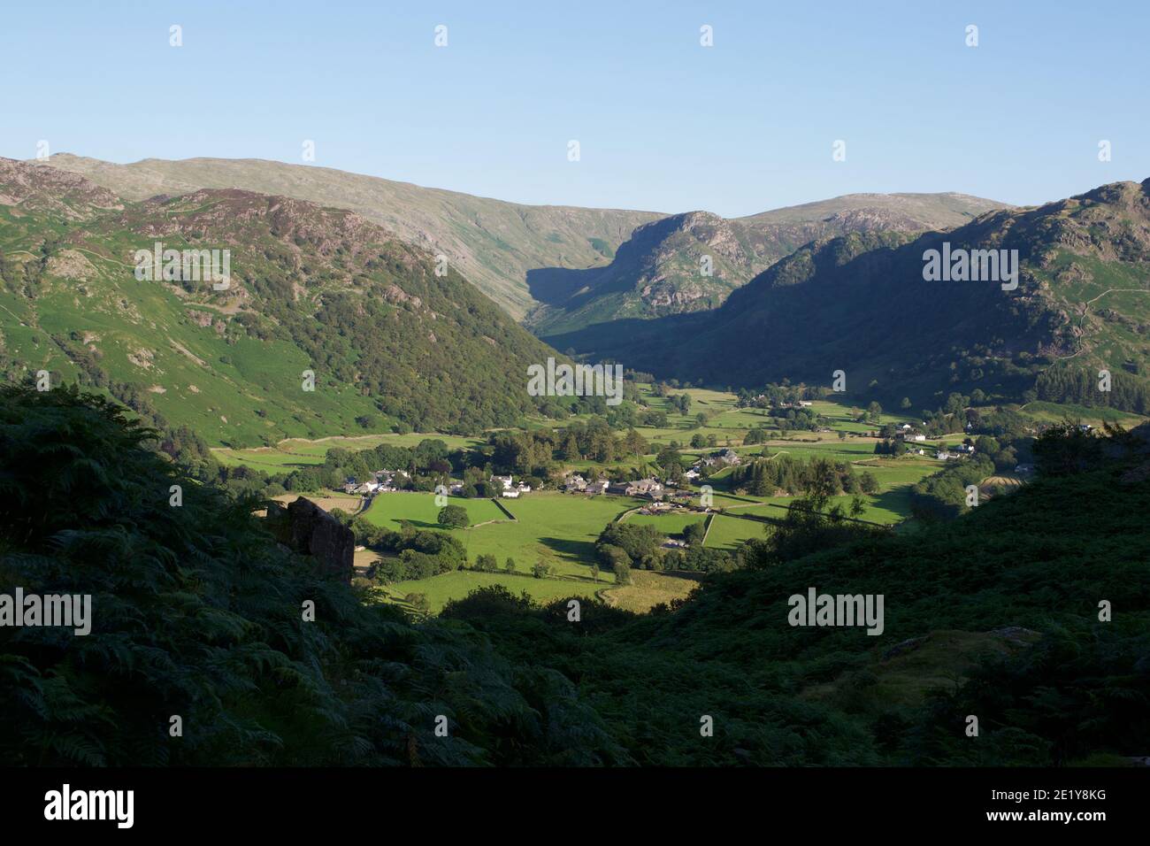 A pastoral valley in the evening sunlight: fields, farms and trees surrounded by high mountains. Long shadows, green bracken and grass in summer time Stock Photo