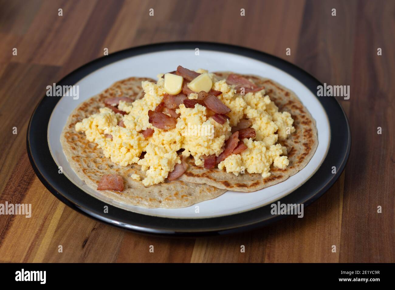 An angled view of Derbyshire oatcakes served with scrambled egg and bacon on a black rimmed plate. Stock Photo