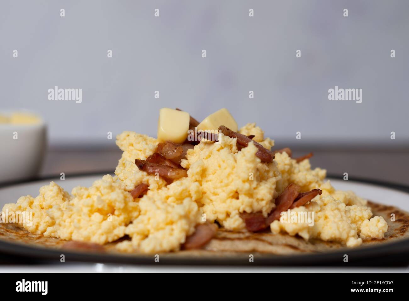 A side view of scrambled eggs and bacon on top of Derbyshire oatcakes sitting on a black rimmed plate. A butter dish is out of focus in the background Stock Photo