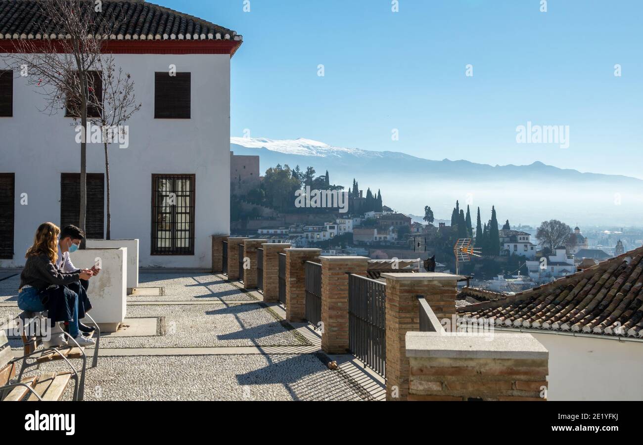 A walk in Granada: Young couple with masks sitting on a bench in the Plaza María Santísima de la Aurora contemplating the city and its monuments Stock Photo