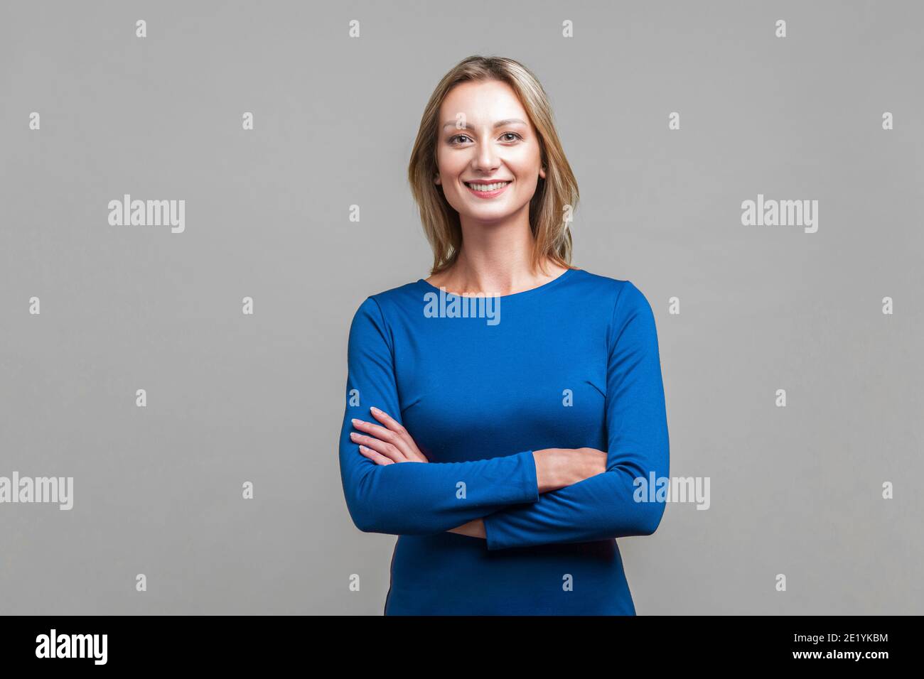 Portrait of happy successful businesswoman in tight blue dress standing with crossed hands, looking at camera with charming and joyous toothy smile. i Stock Photo