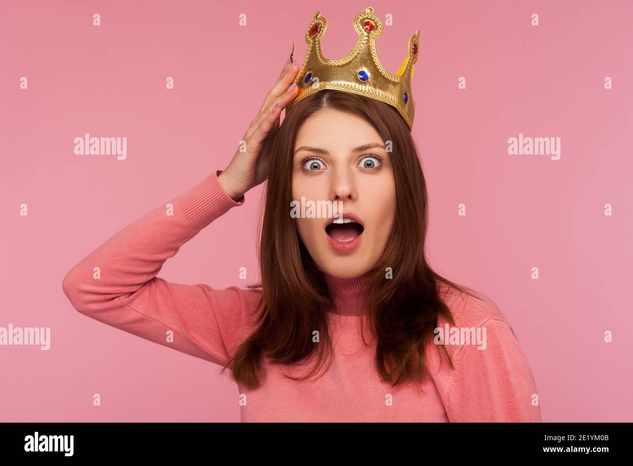 Closeup shocked bossy woman with brown hair in pink sweater adjusting golden crown on head looking at camera with big eyes and open mouth. Indoor stud Stock Photo