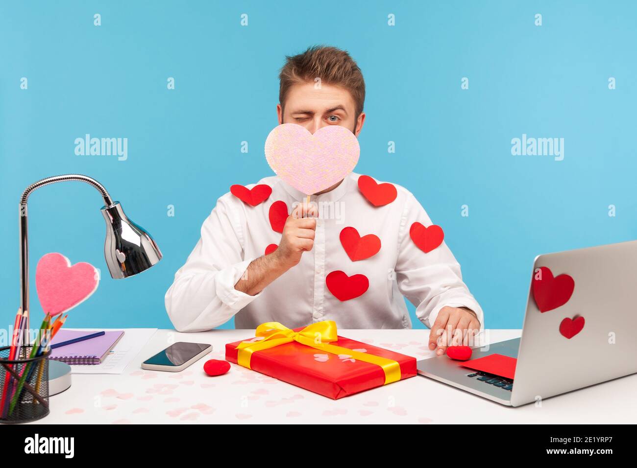 Man office worker in love with hearts on shirt hiding face behind pink heart and winking, preparing gift for valentines day, celebration, romance. Ind Stock Photo