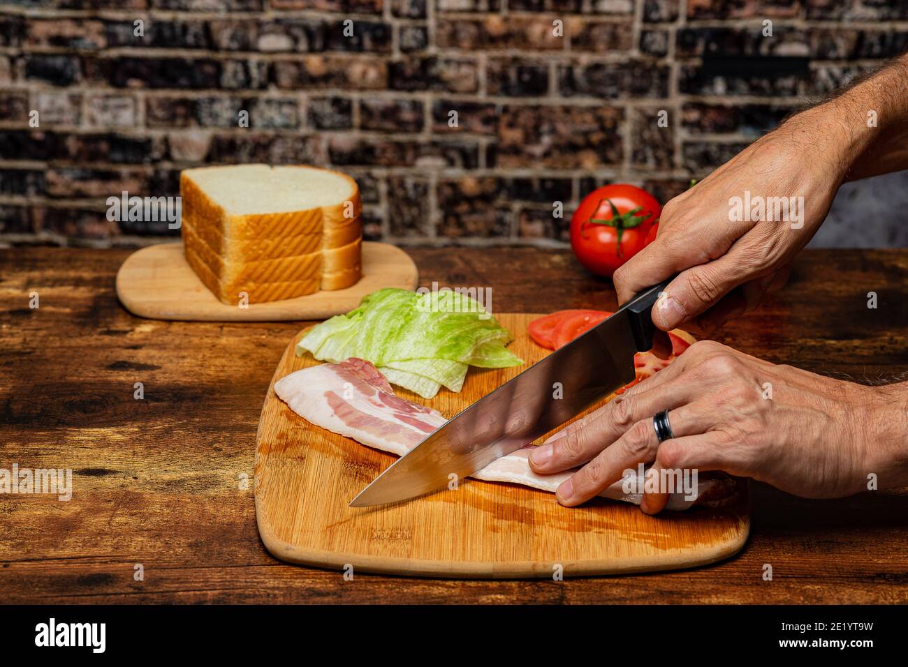 Male Caucasian Hands cutting bacon with a chefs knife on a cutting board and preparing the ingredients for a BLT sandwich. Stock Photo