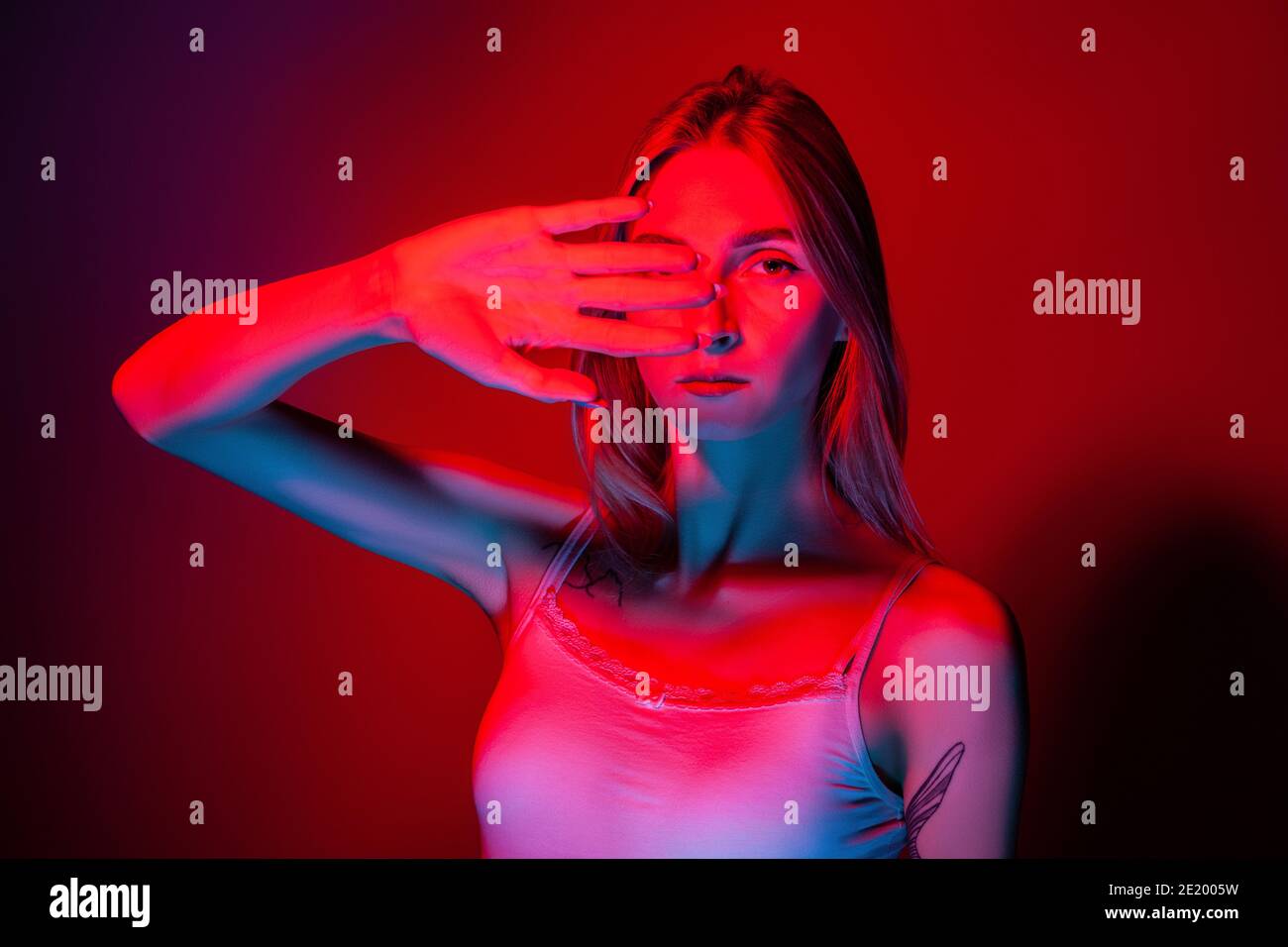 Unemotional young female covering eyes with hand in studio with red light Stock Photo