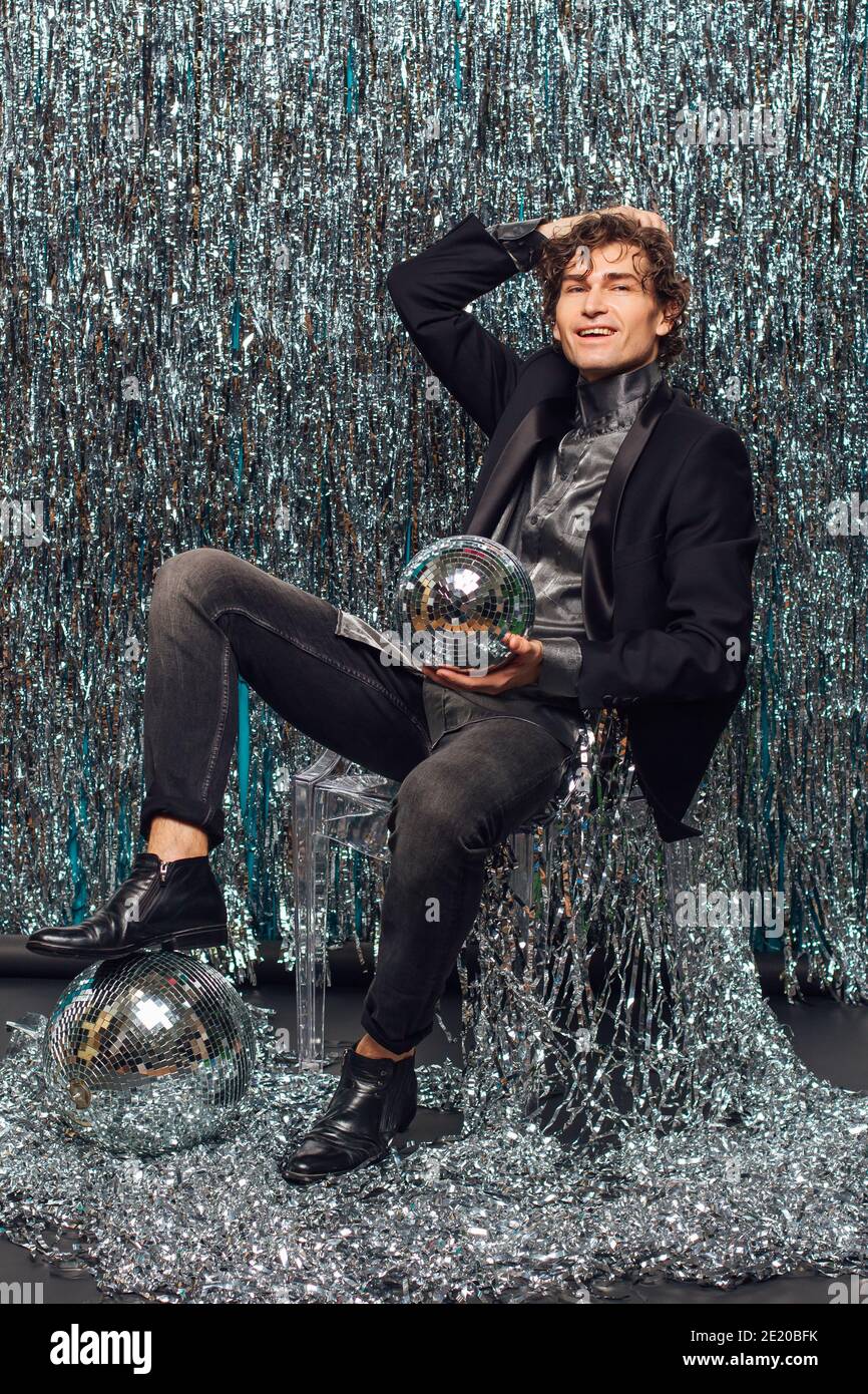 Tall attractive man sitting on the chair over the sparkle background holding mirror disco ball. Stock Photo