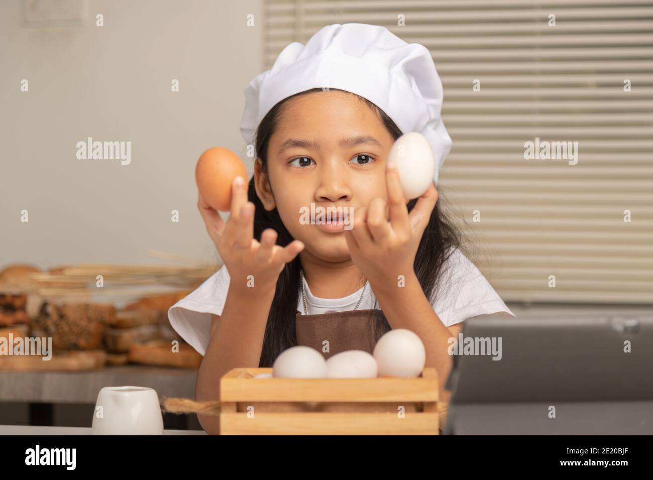 A little Asian girl wearing a white chef hat is choosing between a duck egg and a chicken egg to make a bakery on a white cooking table in the kitchen Stock Photo