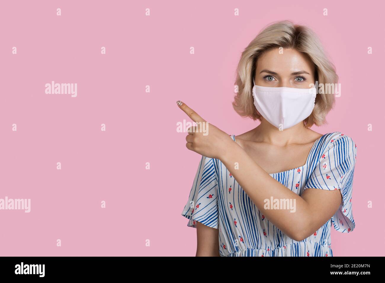 Blonde woman with medical mask is pointing at something on a pink studio wall with free space Stock Photo