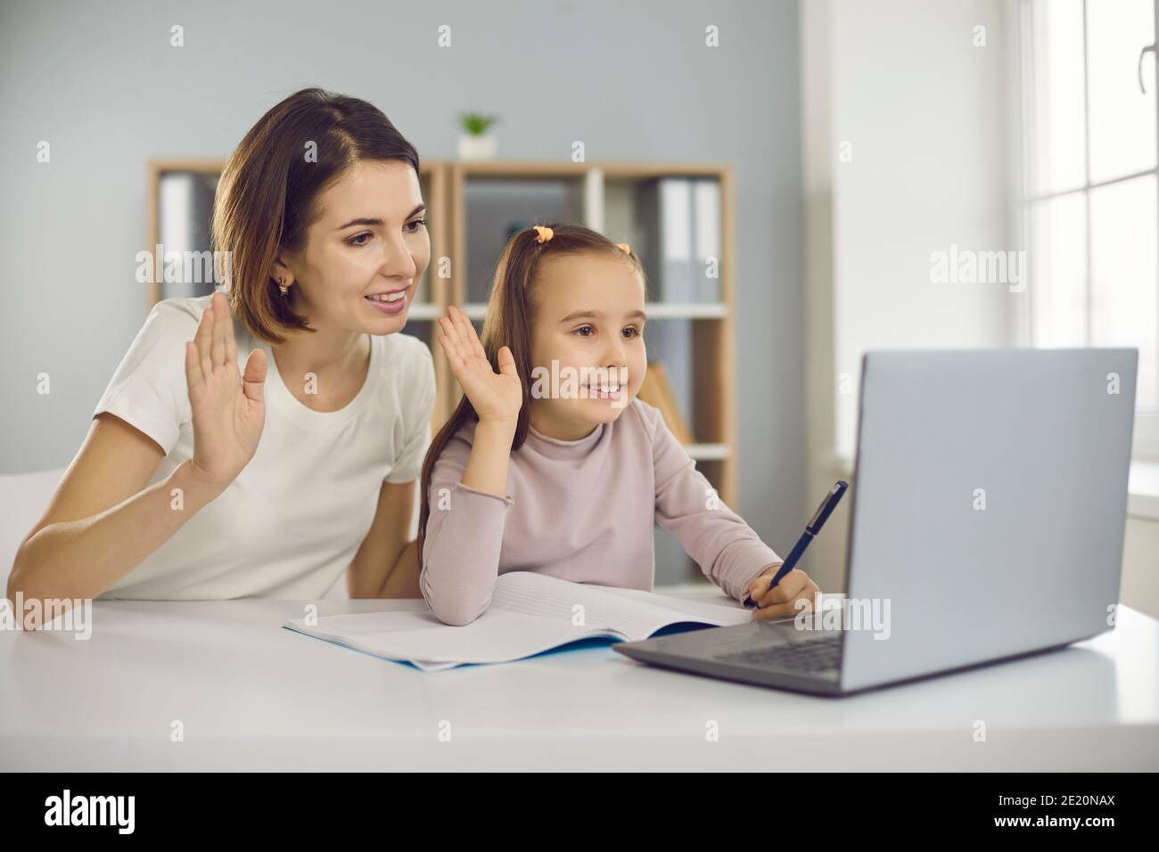 Happy mom and daughter waving hands at laptop, greeting teacher in online lesson Stock Photo