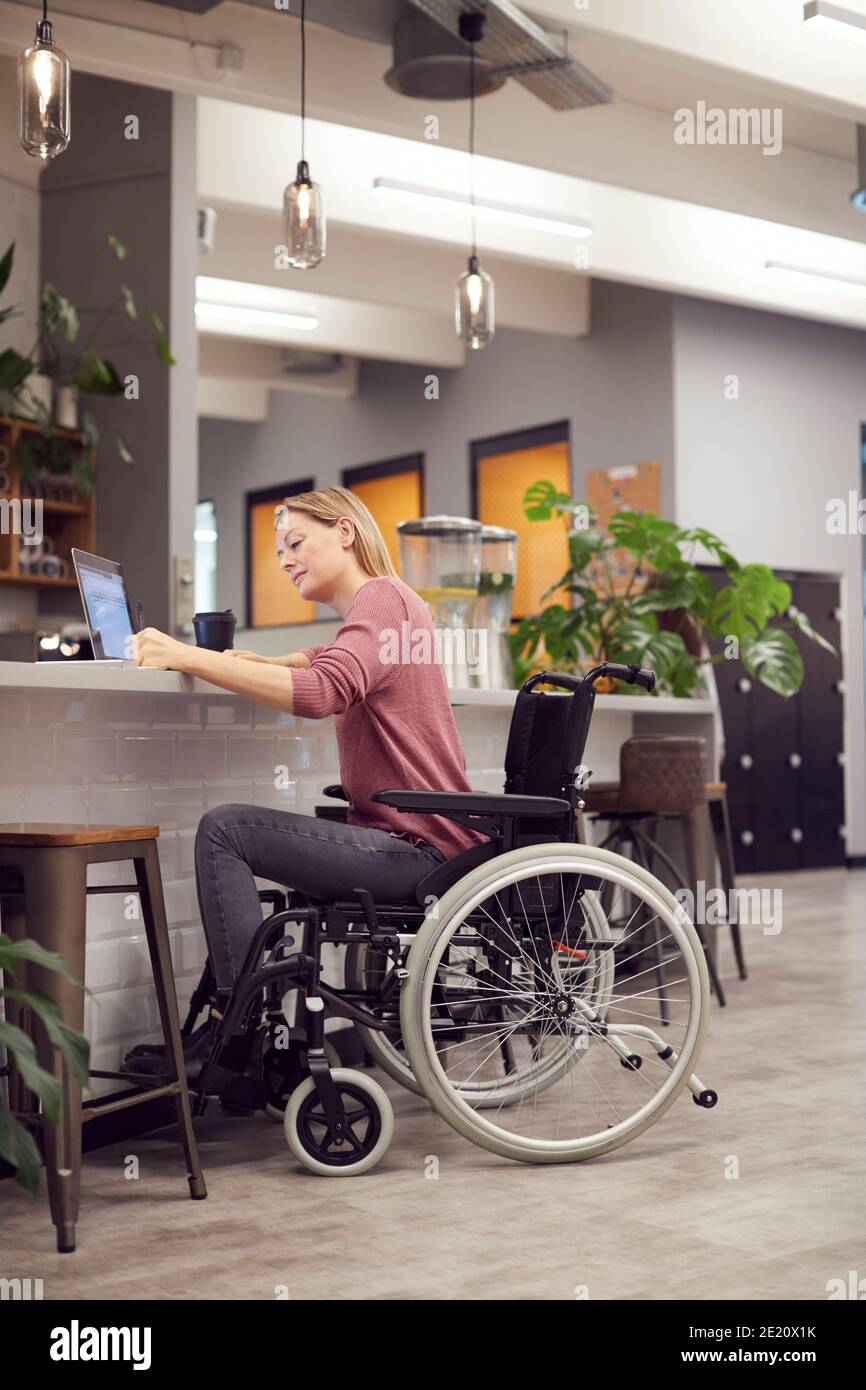 Businesswoman In Wheelchair Working On Laptop In Kitchen Area Of Busy Modern Office Stock Photo