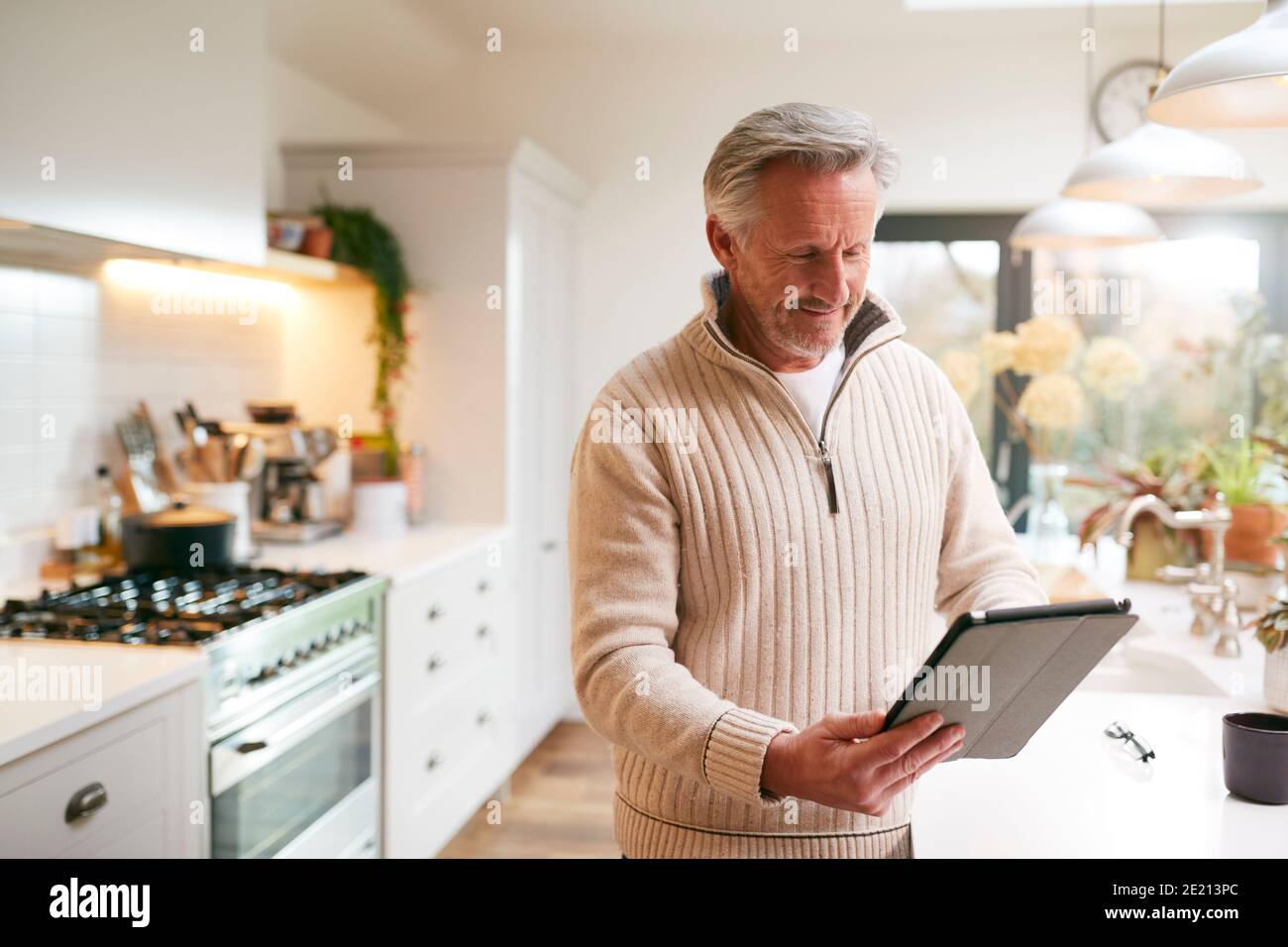 Mature Man In Kitchen Working From Home Using Digital Tablet Stock Photo