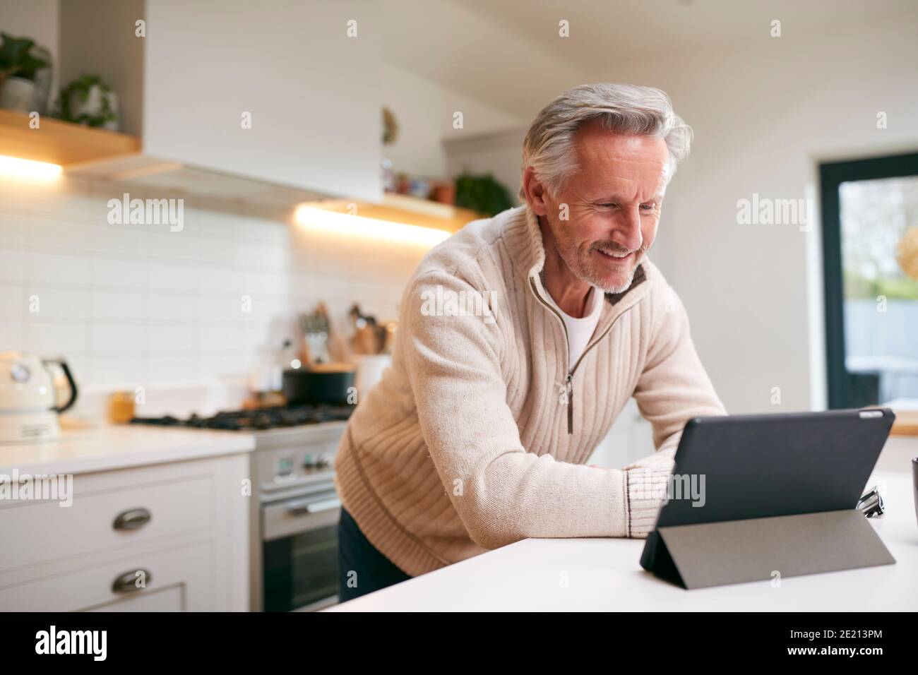 Mature Man In Kitchen Working From Home Using Digital Tablet Stock Photo