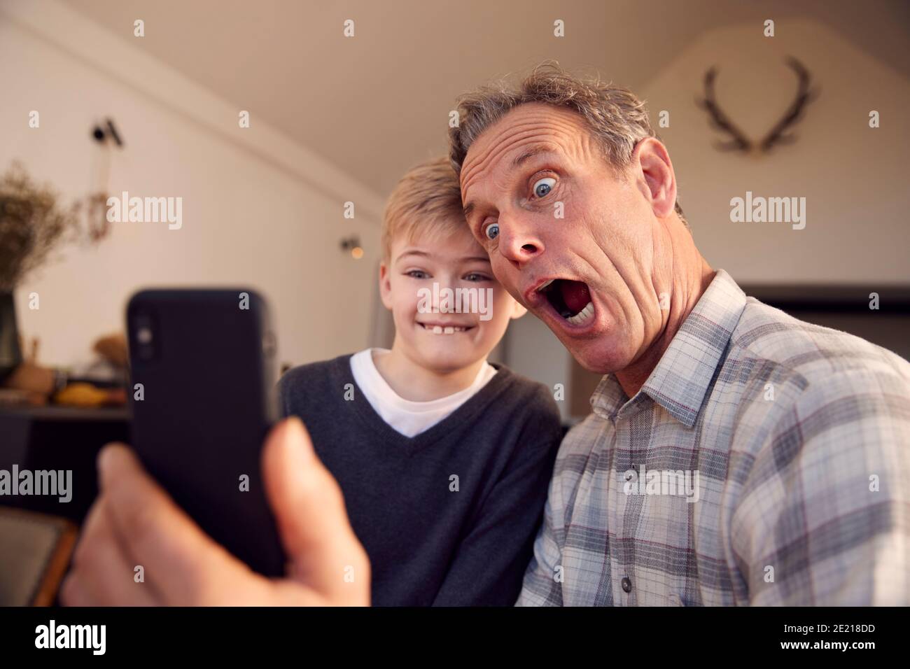 Grandson With Grandfather Pulling Faces And Taking Selfie On Mobile Phone At Home Stock Photo