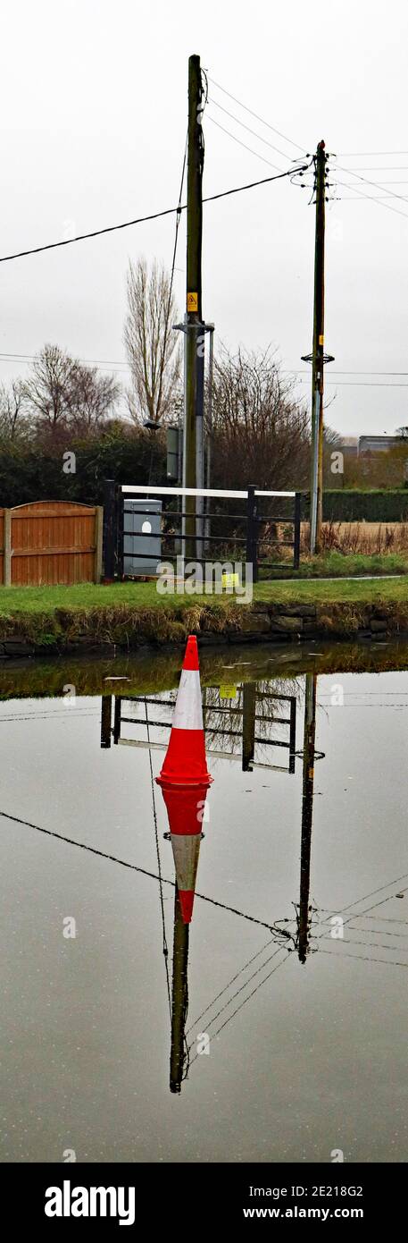 Cone on the ice, water’s of the canal had frozen over, a traffic cone sits on the ice lined up with electric wire poles causing a vertical reflection Stock Photo