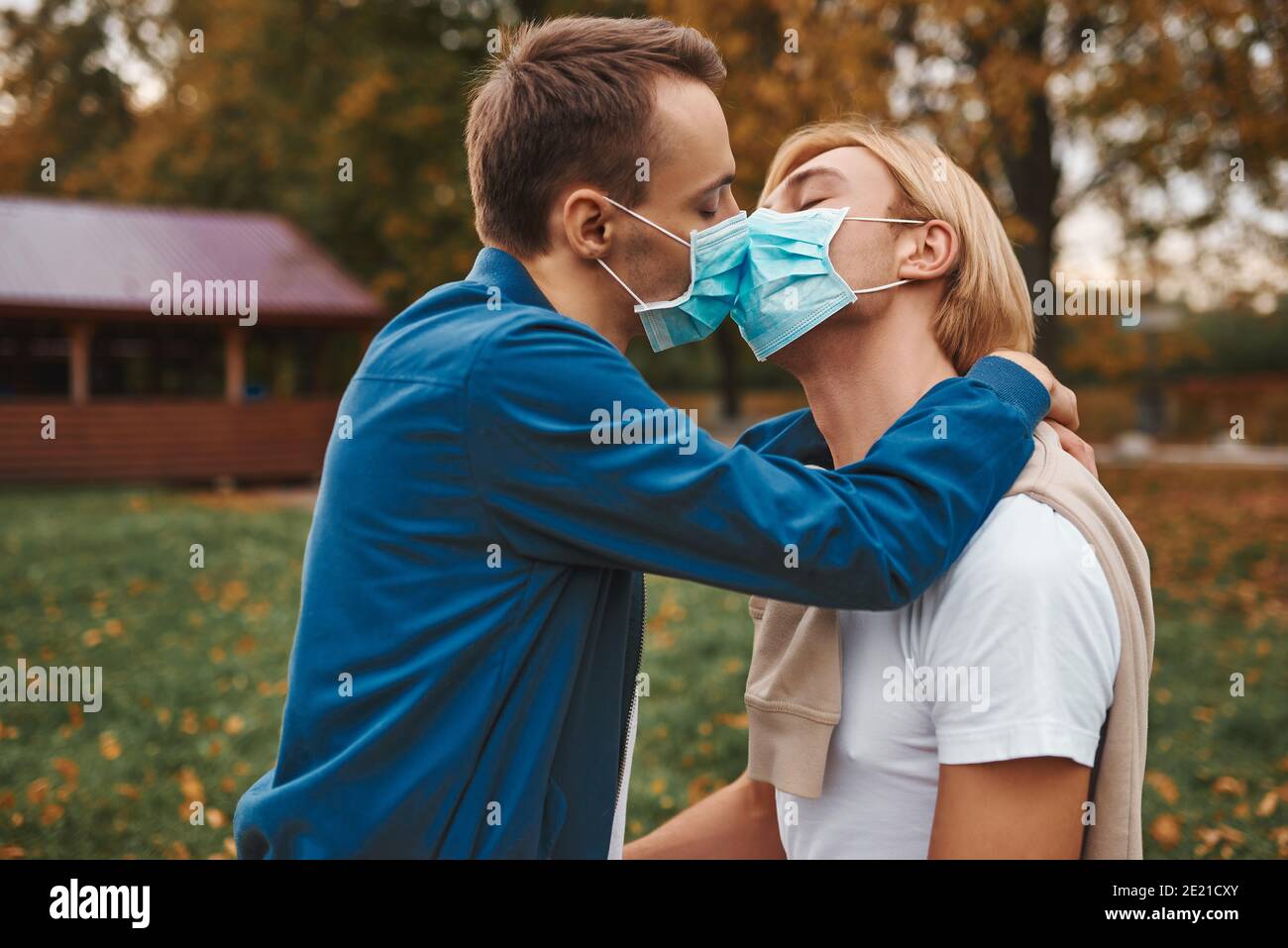 Romantic gay couple outdoors. Love during coronavirus. Two handsome men kissing wearing face masks. Covid-19 protection. Stock Photo