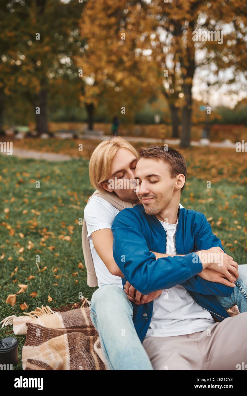 Loving gay couple having romantic date outdoors. Two handsome men sitting together on blanket in park and hugging. LGBT concept. Stock Photo