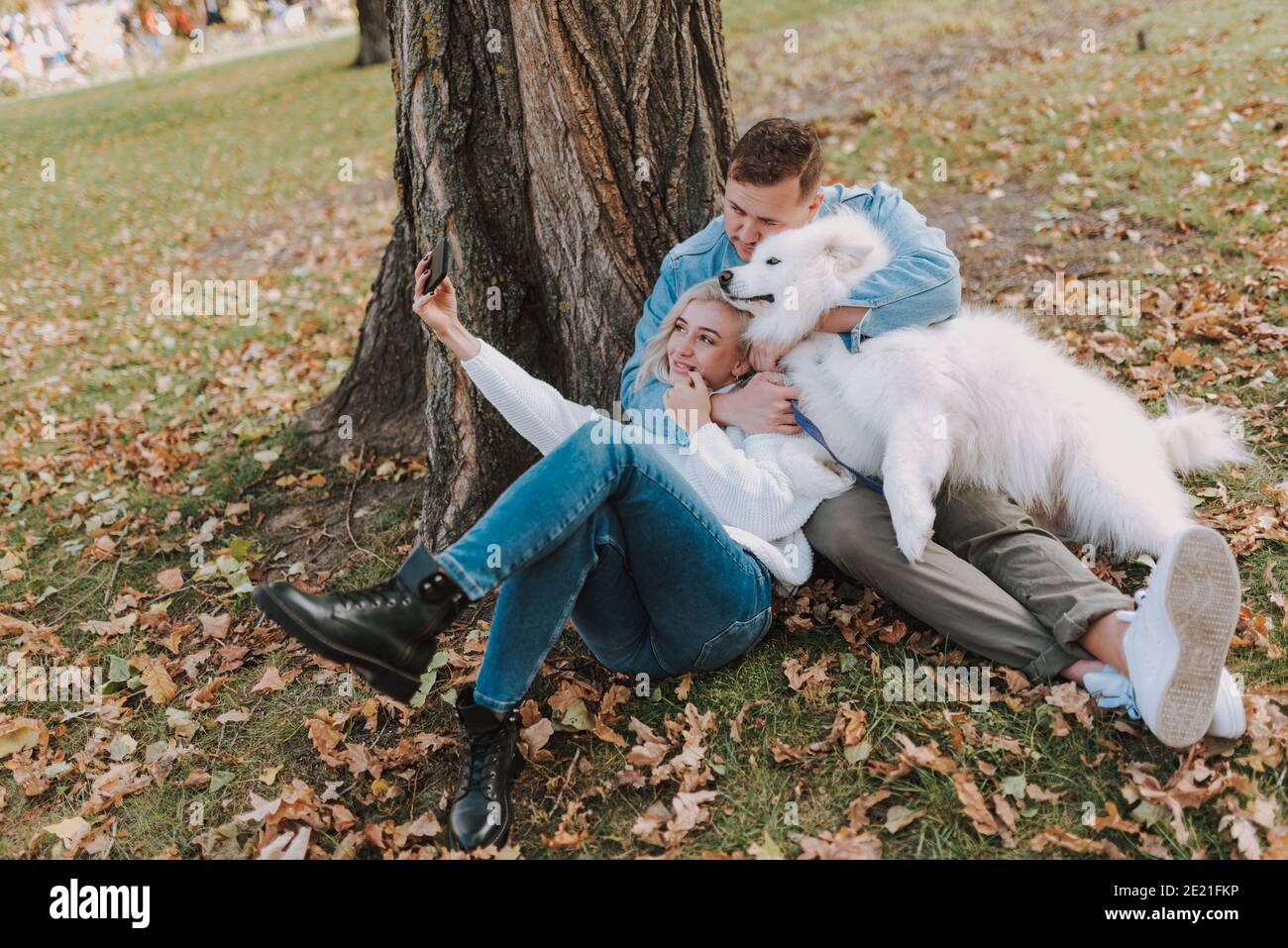 Smiling young couple with dog sitting in the park while making photo on ...