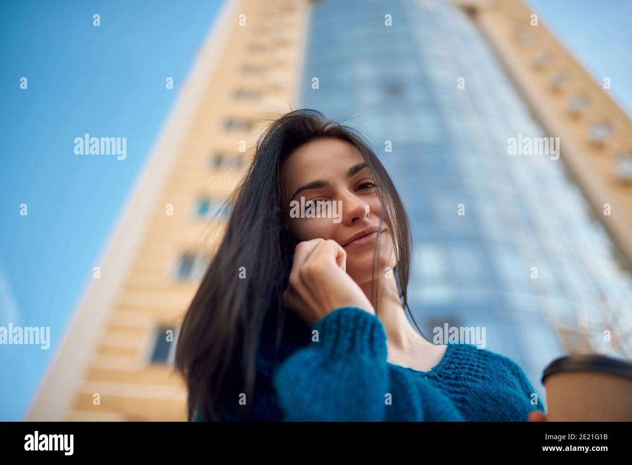 Close up shot of a long-haired woman speaking by cellphone on the street in a good mood, tall office tower is seeing on blurred background Stock Photo