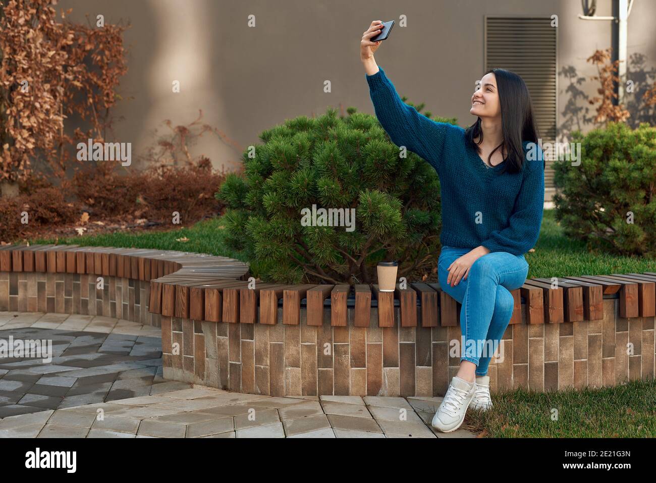 Cheerful young lady in casual clothing making selfie on her smartphone sitting outdoors Stock Photo