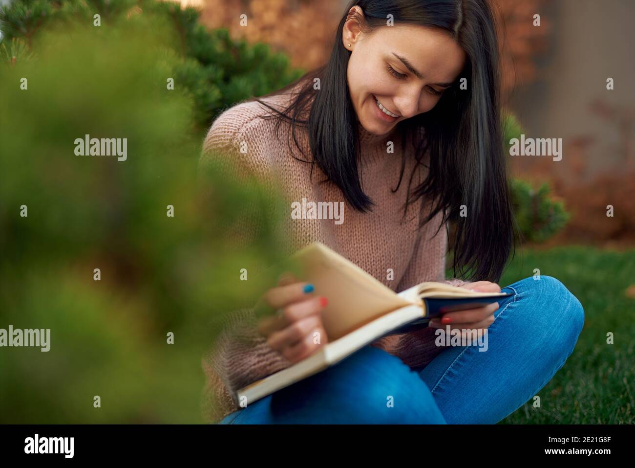 Nice-looking lady sitting cross-legged on fresh green grass outdoors, flipping pages of her workbook with interest Stock Photo