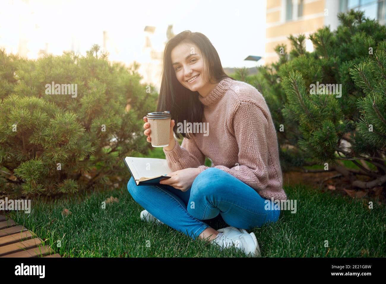 Cheerful young lady with paper glass of coffee in hands holding her memo book on knees while sitting on a cosy green lawn Stock Photo